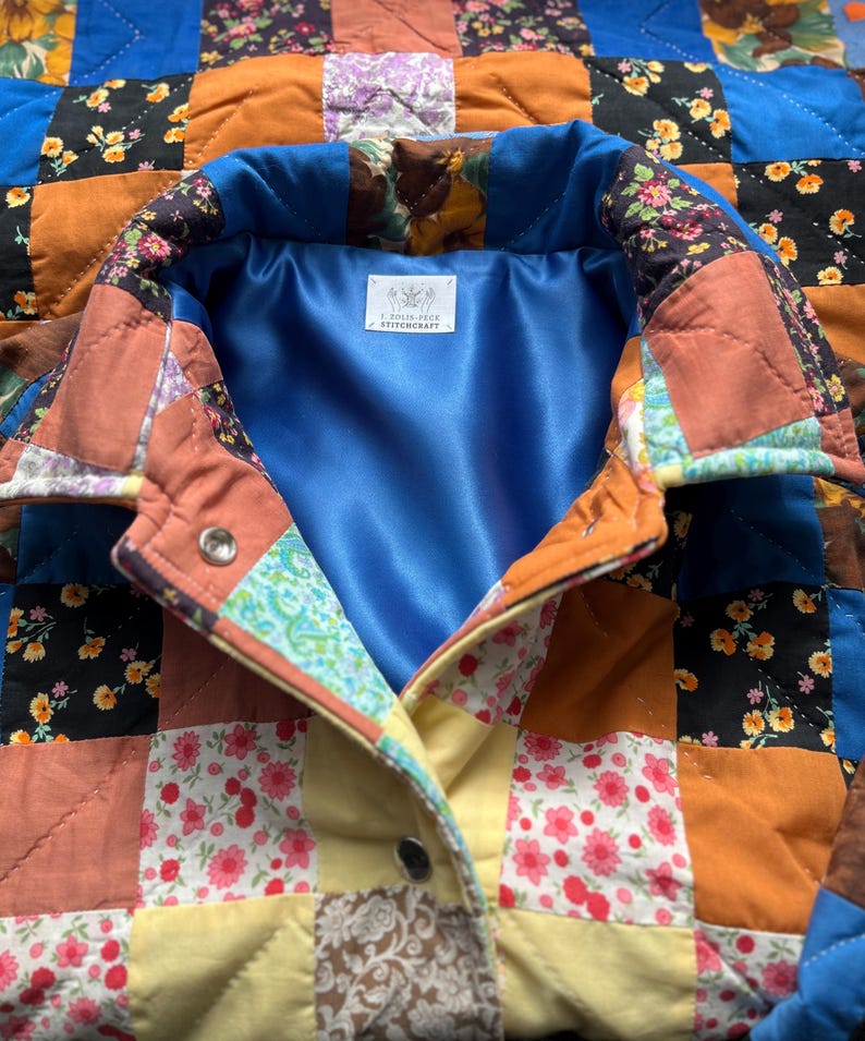 A colourful quilted jacket is displayed on a teal mannequin.  It features a patchwork design with various floral & patterned squares in shades of blue, pink, yellow, orange, and green.  Blue trim outlines the pockets and edges.