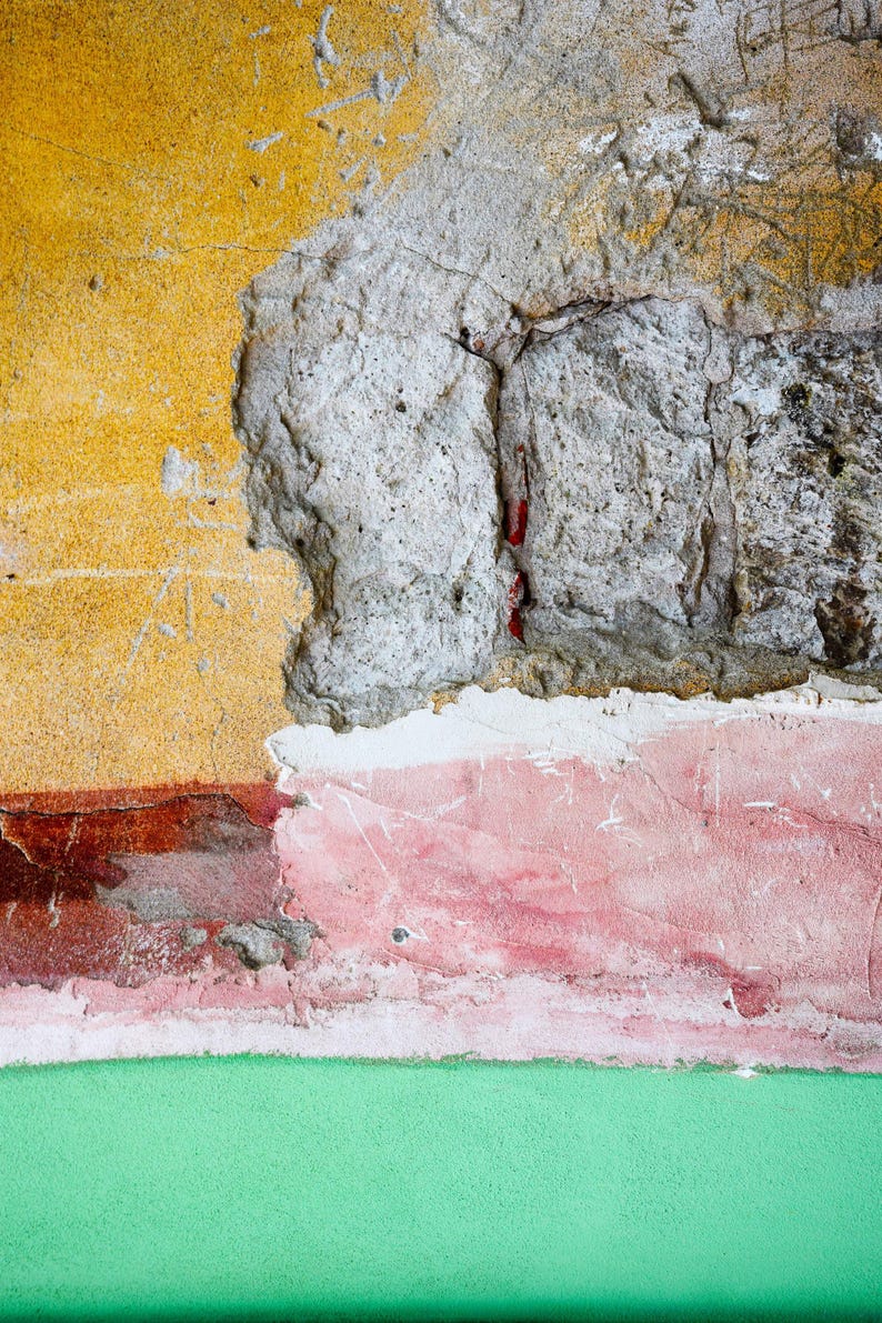 May include: Close-up of a weathered wall with layers of color. The top section is yellow, transitioning to a gray, textured area. Below, a band of red and pink paint meets a green base, creating a textured, abstract composition.