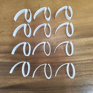 Peut inclure: Une collection de douze formes blanches, abstraites et courbes, peut-être des emporte-pièces, disposées sur une surface en bois. Chaque forme a un design en boucle avec une extension incurvée.