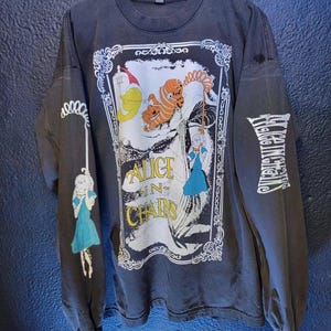May include: Dark gray long-sleeve shirt featuring a graphic from Alice in Chains. The design includes illustrations of characters and the band's name. The left sleeve has a drawing of a girl in a blue dress. The right sleeve has the band's name.