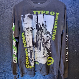 Może przedstawiać: Szara, długorękawowa koszulka z czarno-białym nadrukiem graficznym przedstawiającym zespół. Nazwa zespołu, "Type O Negative", jest nadrukowana białymi literami na czarnym tle. Koszulka ma również napis "World Coming Down" nadrukowany na lewym rękawie zielonymi literami.