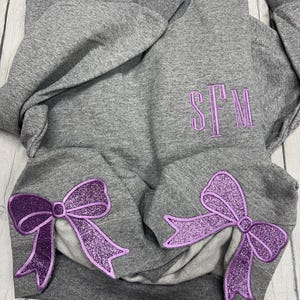 May include: A heather gray crewneck sweatshirt featuring a purple embroidered monogram with the letters SFM on the upper left chest. The lower portion of the sleeves displays two large, glittery purple bow appliques with stitched outlines.