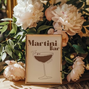Op de afbeelding: Een gouden ingelijst bord met de tekst "Martini Bar" in bruine letters. Onder de tekst staat een bruine lijntekening van een martini-glas met olijven. Het bord staat op een tafel met witte pioenen.