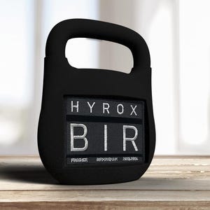 May include: Black kettlebell with a rectangular patch displaying "HYROX BIR" in white lettering. Below, it reads "FINISHER BIRMINGHAM 2023.2024". The kettlebell is on a wooden surface, with a blurred background.