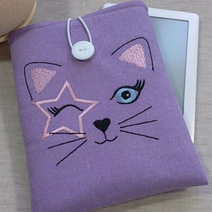 Könnte beinhalten: Eine lavendelfarbene Stoffhülle für einen E-Reader mit einem gestickten Katzengesicht. Die Katze hat ein zwinkerndes Auge, einen Stern und Schnurrhaare. Ein weißer Knopf- und Schlaufenverschluss sichert die Hülle. Ein E-Reader ist teilweise hinter der Hülle sichtbar.