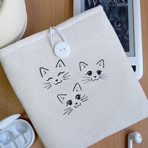 Puede incluir: Una funda de libro electrónico de color crema con tres caras de gato negro bordadas en la parte delantera. La funda tiene un cierre de botón blanco y un lazo. Se ve un lector de libros electrónicos con el título "Warriors".