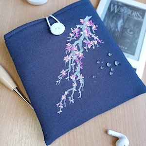 May include: A navy blue e-reader case featuring a floral embroidery design in pink and light blue. The case has a white button closure and a matching loop. The cover is on a wooden surface.