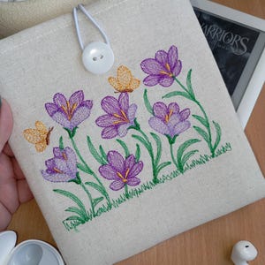 Puede incluir: Funda de tela color crema con bordados de flores de azafrán moradas y mariposas naranjas. La funda tiene un cierre de botón blanco y está diseñada para guardar un lector electrónico. Hierba y hojas verdes bordadas en la base de las flores.