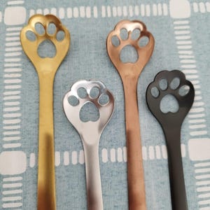 May include: Four stainless steel spoons with a paw print design. The spoons are gold, silver, copper, and black.