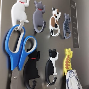 Könnte beinhalten: Eine Sammlung von sieben katzenförmigen Magnethaken in verschiedenen Farben, darunter Weiß, Grau, Schildpatt, getigert, Schwarz sowie Schwarz-Weiß. Jeder Haken zeigt eine Katze mit einem geschwungenen Schwanz als Haken, an dem eine blaue Schere, Schlüssel und ein gemusterter Stoff hängen.