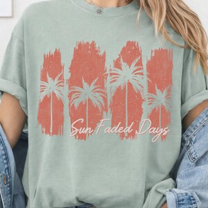 May include: A light green t-shirt featuring a distressed coral-colored graphic of palm trees and the text "Sun Faded Days." The design has a vintage, worn look. The shirt has rolled-up sleeves and a relaxed fit.