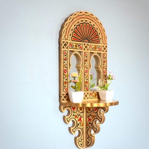 Puede incluir: Estantería de pared decorativa de madera con espejo arqueado, con intrincados patrones florales y geométricos en rojo, amarillo y negro. La estantería contiene dos pequeñas macetas blancas con flores artificiales. El diseño recuerda a la arquitectura marroquí.