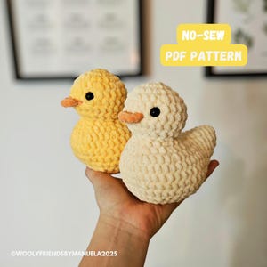 Può includere: Due giocattoli di peluche a forma di anatra all'uncinetto, uno giallo e uno bianco, sono tenuti in una mano. Il testo "NO-SEW PDF PATTERN" è visualizzato sopra le anatre.
