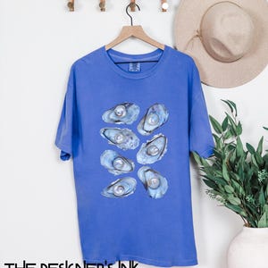 Könnte beinhalten: Ein königsblaues T-Shirt mit einem Aufdruck von acht perlenbesetzten Austern. Die Austern sind in einer Traube angeordnet, wobei die Perlen nach außen zeigen.