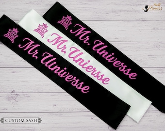 May include: Three satin sashes in black and white, embroidered with pink text and a crown design. The text reads "Mr. Universe" and "Mr. Universe". The sashes are displayed on a light wood grain surface. The words "Custom Sash" are at the bottom.