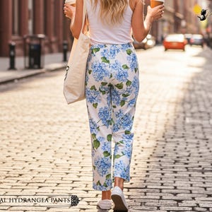 May include: White pants with a blue hydrangea floral print. The pants are straight-legged and are worn with white sneakers. The text "FLORAL HYDRANGEA PANTS" is at the bottom of the image.