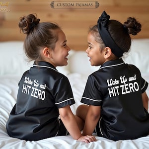 May include: Two children wearing black satin pajamas with white trim and the text "Wake Up and HIT ZERO" on the back. The pajamas are a short-sleeved style.