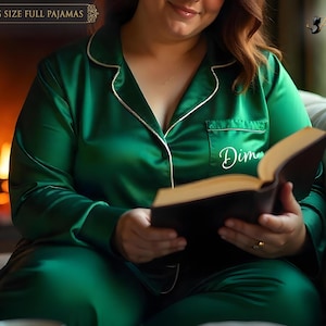 May include: Emerald green plus size full pajamas with white piping. The pajamas include a button-down top with a breast pocket and the name "Dim" embroidered on it. The person is holding a book with a dark cover.