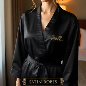 May include: Black satin robe with a personalized embroidered name "Bella" on the left side. The robe has a tie belt and long sleeves.