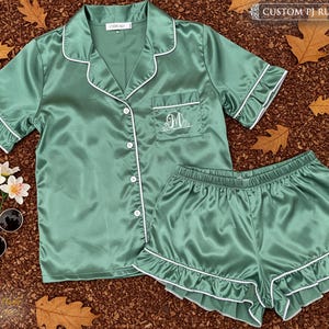 May include: A jade green satin pajama set with white trim, featuring a short-sleeved button-up shirt and matching shorts. The shirt has a pocket with an embroidered initial. The set is labeled "CUSTOM PJ RUFFLE".