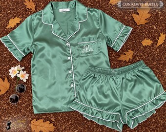 May include: A jade green satin pajama set with white trim, featuring a short-sleeved button-up shirt and matching shorts. The shirt has a pocket with an embroidered initial. The set is labeled "CUSTOM PJ RUFFLE".