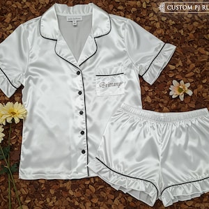 May include: White satin pajama set with black trim, featuring a short-sleeved button-down shirt and matching shorts. The shirt has a breast pocket embroidered with "Brittany". The set is displayed on a brown surface with flowers. Text reads "CUSTOM PJ RUFFLE".