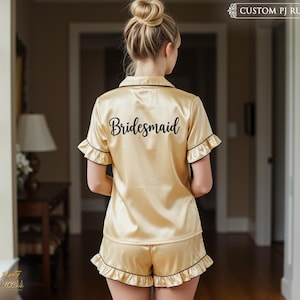 May include: A gold satin pajama set with black trim and the word "bridesmaid" embroidered on the back. The set includes a short-sleeved top and shorts.