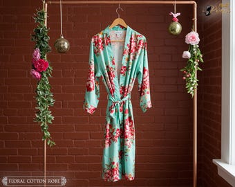 May include: A floral cotton robe in a light teal color with a red floral pattern. The robe is hanging on a wooden hanger and tied with a matching belt. The robe is displayed on a copper clothing rack.