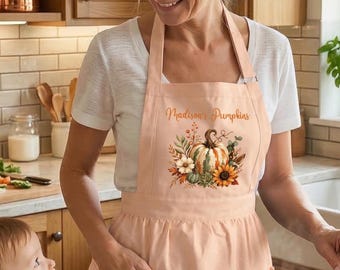 Puede incluir: Un delantal de color melocotón claro con un gráfico central de una calabaza rodeada de flores y follaje de otoño, con el texto 'Madison's Pumpkins' impreso sobre el diseño. El delantal incluye dos bolsillos con volantes en el frente.