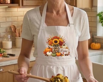 Puede incluir: Un delantal blanco con un gráfico otoñal colorido de girasoles, calabazas y un letrero que dice Violet. El delantal se lleva sobre una camiseta blanca, con un cuenco de madera lleno de relleno y un pastel bundt sobre la encimera.