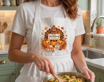 Puede incluir: Un delantal blanco con un gráfico colorido de Acción de Gracias que muestra un pavo, calabazas y el texto Happy Thanksgiving y Evelyn, usado sobre una camiseta blanca mientras se mezclan alimentos en un tazón de cobre rodeado de platos.