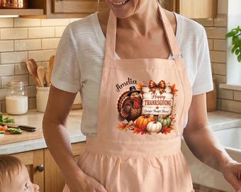 Puede incluir: Un delantal de color melocotón claro con un gráfico de un pavo, calabazas y el texto "Amelia Happy Thanksgiving" impreso en la parte delantera. El delantal tiene dos bolsillos frontales y se lleva sobre una camiseta blanca en una cocina.
