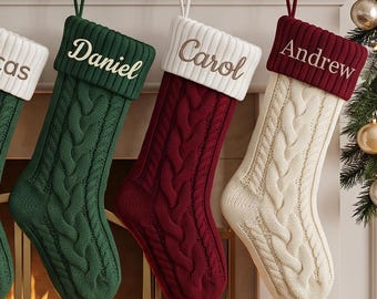 Op de afbeelding: Zes kerstsokken van kabelgebreid materiaal hangen aan een schoorsteenmantel, elk met een omgeslagen boord geborduurd met de namen Amelia, Heather, Lucas, Daniel, Carol en Andrew. De sokken zijn beige, wit, groen, donkergroen, rood en bordeaux.