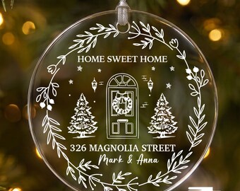 Puede incluir: Un adorno circular transparente de acrílico con diseños blancos grabados de una puerta de entrada, dos pinos y un borde de corona floral. El texto dice HOME SWEET HOME, 326 MAGNOLIA STREET y Mark & Anna. Cuelga de un cordón plateado.