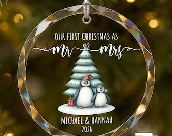 Puede incluir: Un adorno navideño de cristal circular y facetado que cuelga de un cordón plateado. El adorno presenta el texto "OUR FIRST CHRISTMAS AS MR & MRS", "MICHAEL & HANNAH" y "2026" sobre y bajo una ilustración de dos pingüinos junto a un árbol.