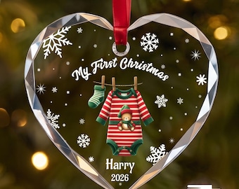 Puede incluir: Un adorno de acrílico transparente en forma de corazón con bordes facetados, colgado de una cinta roja. Presenta copos de nieve blancos, el texto "My First Christmas", un gráfico de un body a rayas con un osito y el nombre "Harry 2026".