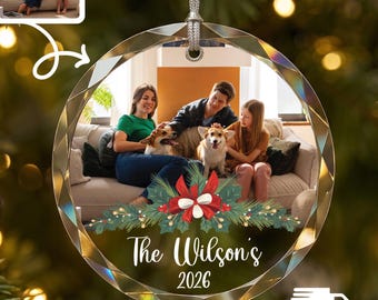 Puede incluir: Un adorno navideño de cristal redondo y facetado con una fotografía impresa de una familia con dos perros en un sofá. El adorno incluye una ilustración festiva de ramas de pino y un lazo rojo, con el texto The Wilson's y 2026 impreso debajo.