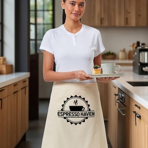 May include: A beige apron with the text "Espresso Haven" in a black circular design. The apron is worn by a person holding a tray with a cup of coffee and a small pastry. The person is in a kitchen setting.