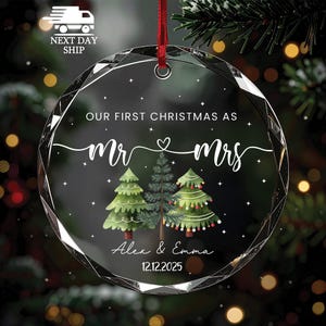 May include: A clear, faceted glass Christmas ornament with a red ribbon. The ornament features the text "OUR FIRST CHRISTMAS AS Mr & Mrs" with a heart symbol, three illustrated Christmas trees, and the names "Alex & Emma" with the date "12.12.2025".