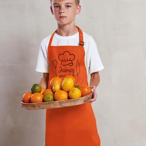 May include: An orange apron with a chef hat graphic, whisk, rolling pin, and the name "James" in script. The apron is worn by a person holding a wooden tray filled with oranges, limes, and lemons. The person is wearing a white t-shirt.