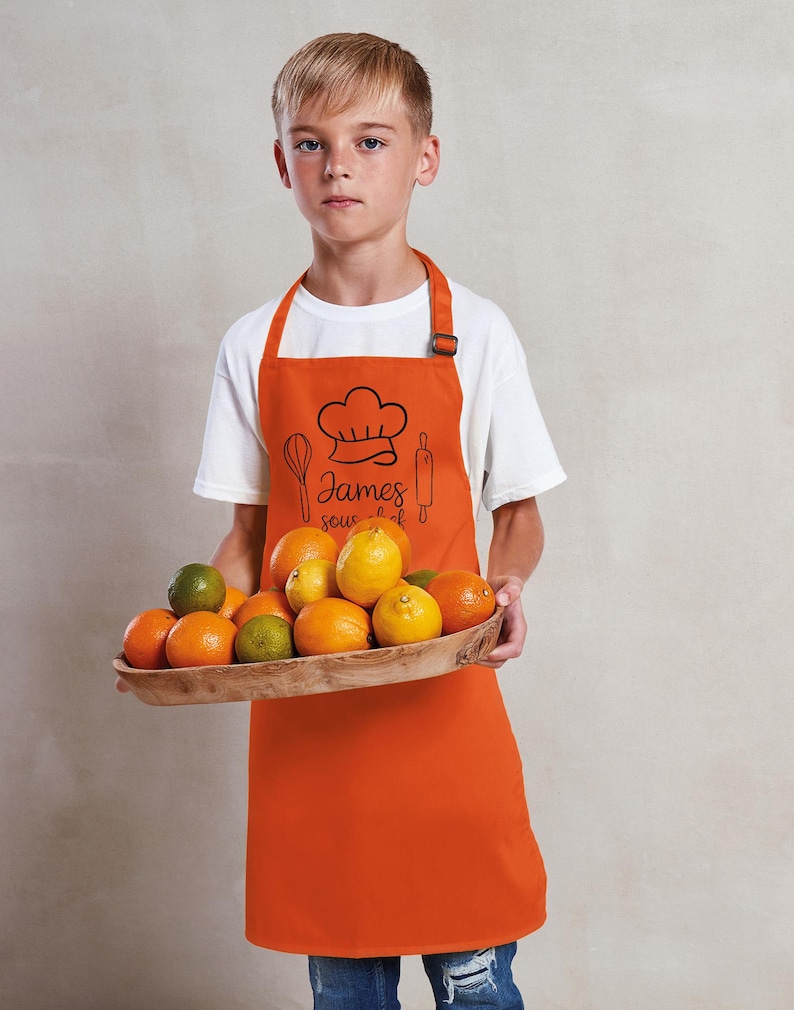 May include: An orange apron with a chef hat graphic, whisk, rolling pin, and the name "James" in script. The apron is worn by a person holding a wooden tray filled with oranges, limes, and lemons. The person is wearing a white t-shirt.