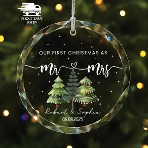 May include: A clear, round Christmas ornament with a silver hanging loop. The ornament features the text "OUR FIRST CHRISTMAS AS Mr & Mrs" with a heart symbol, along with the names "Robert & Sophia" and the date "08.08.2025". Three illustrated Christmas trees are also included.