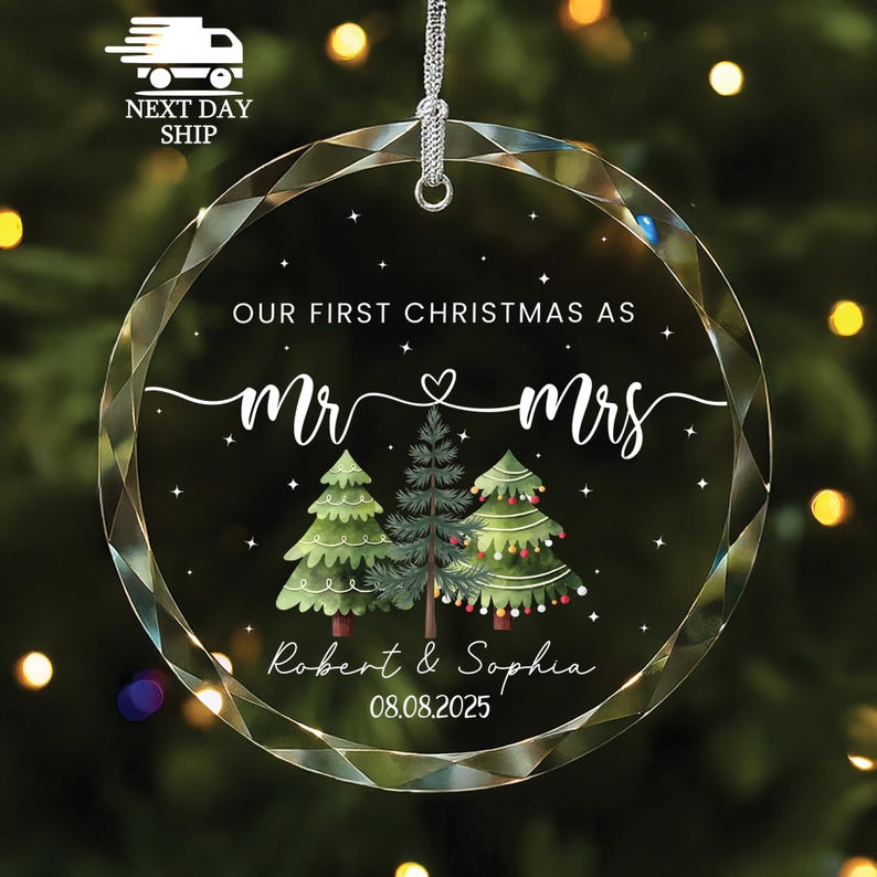 May include: A clear, round Christmas ornament with a silver hanging loop. The ornament features the text "OUR FIRST CHRISTMAS AS Mr & Mrs" with a heart symbol, along with the names "Robert & Sophia" and the date "08.08.2025". Three illustrated Christmas trees are also included.
