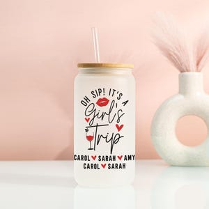 May include: Frosted glass tumbler with a bamboo lid and straw. The tumbler features the text "OH SIP! IT'S A Girl's Trip" in black and red, with names below. A decorative vase and heart-shaped sign are in the background.