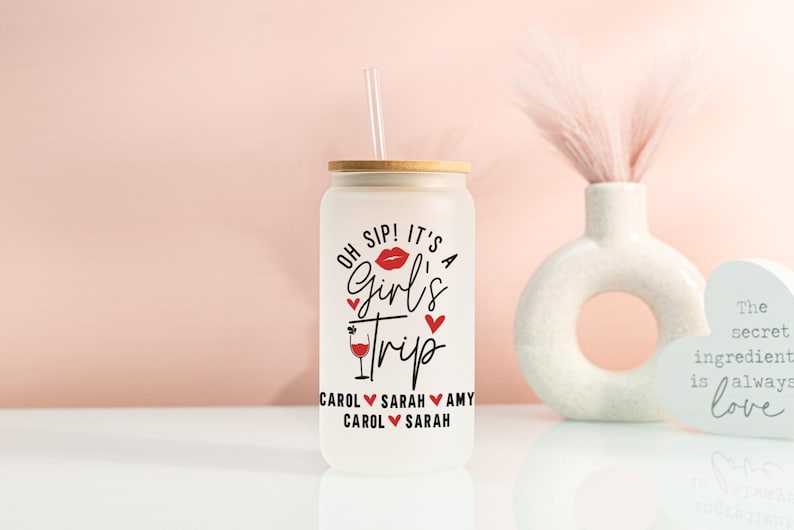 May include: Frosted glass tumbler with a bamboo lid and straw. The tumbler features the text "OH SIP! IT'S A Girl's Trip" in black and red, with names below. A decorative vase and heart-shaped sign are in the background.
