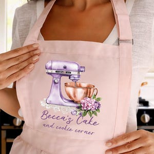 May include: A light pink apron featuring a purple stand mixer with a copper bowl, a floral arrangement, and the text "Becca's Cake and cookie corner". The apron is made of a soft material and has adjustable straps.