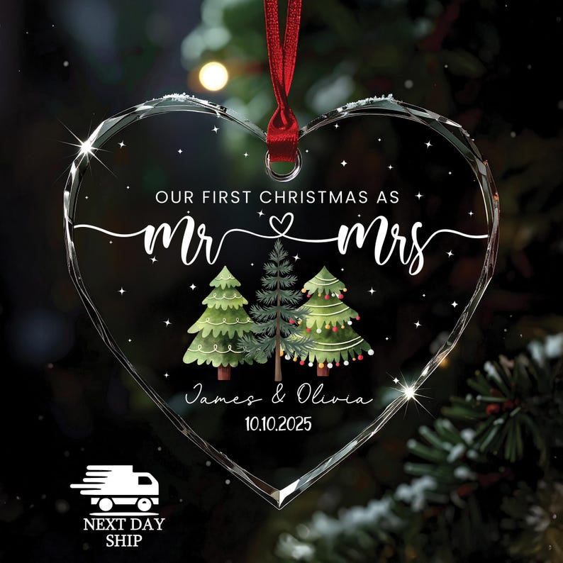 May include: Heart-shaped clear glass Christmas ornament with a red ribbon. The ornament features the text "OUR FIRST CHRISTMAS AS Mr & Mrs" and the names "James & Olivia" with the date "10.10.2025". Three illustrated Christmas trees are also featured.