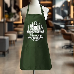 May include: A dark green apron with a white floral design and the name "Thomas" in a script font. The design includes nail care tools. The apron has two front pockets and an adjustable neck strap.