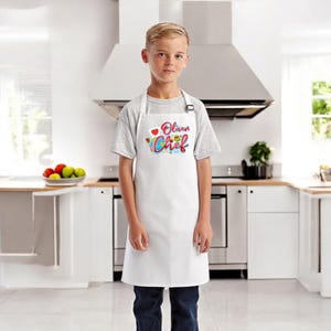 May include: A white apron with the text "Oliver Chef" in colorful lettering, worn by a person. The apron has a heart and a chef's hat graphic. The person is wearing a gray t-shirt and blue jeans, standing in a kitchen.