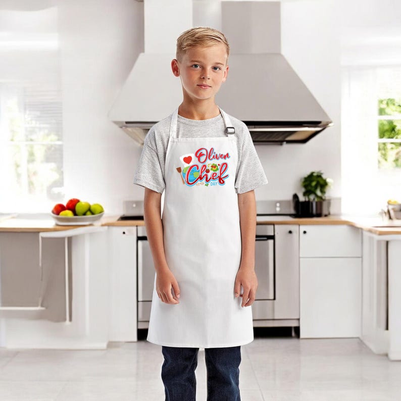 May include: A white apron with the text "Oliver Chef" in colorful lettering, worn by a person. The apron has a heart and a chef's hat graphic. The person is wearing a gray t-shirt and blue jeans, standing in a kitchen.
